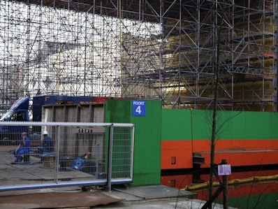 A construction site featuring extensive scaffolding with a bright green and orange barrier labeled 'Poort 4'. Two construction workers, one in overalls and the other wearing a helmet, sit on concrete blocks behind a fence. A blue truck is partially visible in the background.