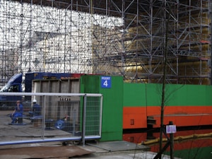 A construction site featuring extensive scaffolding with a bright green and orange barrier labeled 'Poort 4'. Two construction workers, one in overalls and the other wearing a helmet, sit on concrete blocks behind a fence. A blue truck is partially visible in the background.