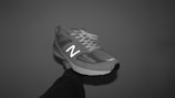 A single athletic shoe is held by a hand against a plain background. The shoe has a mesh upper with synthetic overlays and features a prominent N logo on the side. It has laces tied at the top.