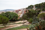 A picturesque park landscape featuring lush greenery and uniquely designed buildings in the background. People are strolling along paved paths surrounded by a variety of trees and shrubs. The scene includes colorful architectural structures with distinctive designs, which add a touch of whimsy to the environment. The horizon shows widespread urban development and distant hills.