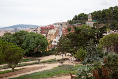 A picturesque park landscape featuring lush greenery and uniquely designed buildings in the background. People are strolling along paved paths surrounded by a variety of trees and shrubs. The scene includes colorful architectural structures with distinctive designs, which add a touch of whimsy to the environment. The horizon shows widespread urban development and distant hills.