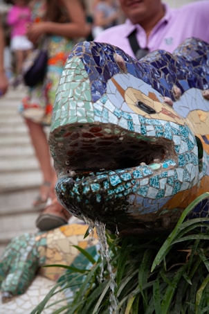 A detailed mosaic lizard sculpture serves as a water fountain with water trickling from its mouth. The sculpture is covered in vibrant tiles featuring a mix of blues, greens, and browns. Surrounding the sculpture are green plants, and there are people in the background, some wearing colorful outfits.
