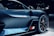 A sleek and modern high-performance sports car is shown in close-up. The image focuses on the car's aerodynamic design with sharp lines and a glossy finish. The vehicle features stylish alloy wheels and a prominent side mirror, with a flag detail on the side skirt.