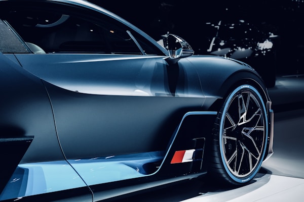 A sleek and modern high-performance sports car is shown in close-up. The image focuses on the car's aerodynamic design with sharp lines and a glossy finish. The vehicle features stylish alloy wheels and a prominent side mirror, with a flag detail on the side skirt.