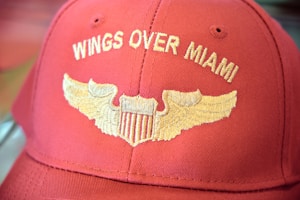 A red baseball cap features gold embroidered wings and the text 'Wings Over Miami.' The stitching includes a stylized emblem resembling a shield flanked by wings.