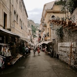 A narrow street lined with traditional European buildings presents a bustling market scene with people walking and shopping. Numerous stalls and shops display various goods, including clothing and artworks, under awnings. The atmosphere is lively with a mix of tourists and locals engaging in the vibrant setting.