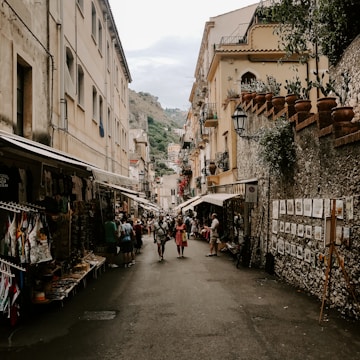 A narrow street lined with traditional European buildings presents a bustling market scene with people walking and shopping. Numerous stalls and shops display various goods, including clothing and artworks, under awnings. The atmosphere is lively with a mix of tourists and locals engaging in the vibrant setting.