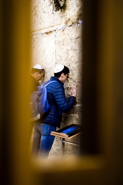 Two individuals wearing kippahs are seen engaged in prayer at an ancient stone wall, with pieces of paper inserted between the stones. One person is resting a hand on the wall, while a wooden stand with books is nearby. The scene is framed by a blurred yellow foreground.