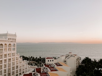 A coastal view featuring a multistory hotel building with arched windows on the left side. The ocean stretches out in the background under a calm, pastel-hued sky transitioning from soft orange to light blue. Palm trees are visible between the building and the water, adding a tropical feel to the scene.