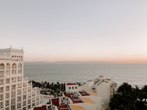 A coastal view featuring a multistory hotel building with arched windows on the left side. The ocean stretches out in the background under a calm, pastel-hued sky transitioning from soft orange to light blue. Palm trees are visible between the building and the water, adding a tropical feel to the scene.