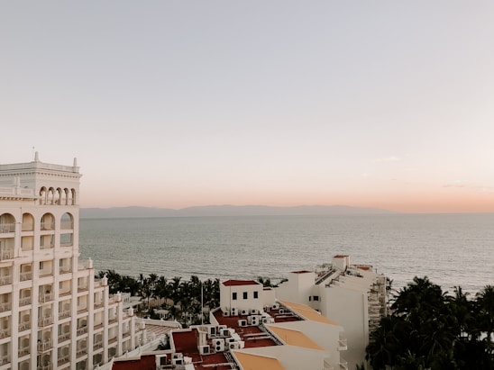 A coastal view featuring a multistory hotel building with arched windows on the left side. The ocean stretches out in the background under a calm, pastel-hued sky transitioning from soft orange to light blue. Palm trees are visible between the building and the water, adding a tropical feel to the scene.