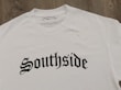 A white T-shirt displayed on a wooden surface with the word 'Southside' printed in bold, gothic-style black lettering. A clothing label reading 'VannyClothingCo.' is visible near the collar, and a blank tag is attached to the shirt.