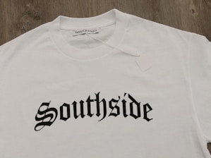 A white T-shirt displayed on a wooden surface with the word 'Southside' printed in bold, gothic-style black lettering. A clothing label reading 'VannyClothingCo.' is visible near the collar, and a blank tag is attached to the shirt.