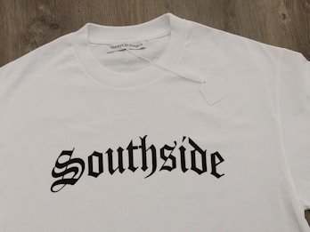 A white T-shirt displayed on a wooden surface with the word 'Southside' printed in bold, gothic-style black lettering. A clothing label reading 'VannyClothingCo.' is visible near the collar, and a blank tag is attached to the shirt.