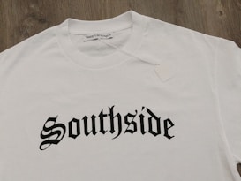 A white T-shirt displayed on a wooden surface with the word 'Southside' printed in bold, gothic-style black lettering. A clothing label reading 'VannyClothingCo.' is visible near the collar, and a blank tag is attached to the shirt.