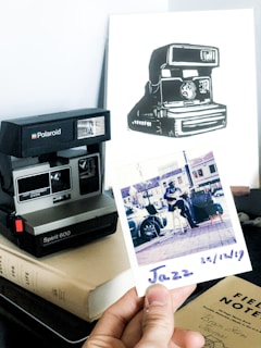 A Polaroid camera is positioned on top of a book, next to a black and white illustration of a Polaroid camera. In the foreground, a hand holds a Polaroid photograph labeled 'Jazz', featuring an outdoor scene with a person playing guitar. Nearby, there is a field notes journal.