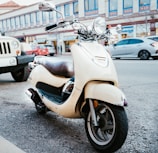 A cream-colored scooter is parked on the side of a busy street. The scooter has a sleek design with a leather seat and chrome accents. Behind it, vehicles including a jeep and a car are part of the urban street scene. The background features a row of shops with large windows and some pedestrians.