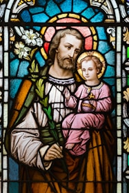 A stained glass artwork depicting a bearded man holding a child. The man is dressed in robes and holds a white lily, while the child is seated in his arm, wearing a pink robe and holding a symbolic object. The background consists of intricate patterns in vibrant colors, including blue and red, forming a halo effect around both figures.
