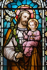 A stained glass artwork depicting a bearded man holding a child. The man is dressed in robes and holds a white lily, while the child is seated in his arm, wearing a pink robe and holding a symbolic object. The background consists of intricate patterns in vibrant colors, including blue and red, forming a halo effect around both figures.