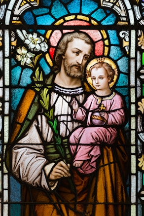A stained glass artwork depicting a bearded man holding a child. The man is dressed in robes and holds a white lily, while the child is seated in his arm, wearing a pink robe and holding a symbolic object. The background consists of intricate patterns in vibrant colors, including blue and red, forming a halo effect around both figures.