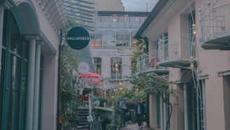 A cozy outdoor shopping area with several small shops and decorative lighting. The alley is flanked by buildings with balconies and glass windows. There are potted plants and greenery along the path, creating a pleasant and inviting atmosphere. A few people are walking through the space, and a prominent sign reads 'HALLHUBER'.