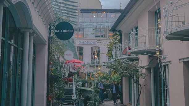 A cozy outdoor shopping area with several small shops and decorative lighting. The alley is flanked by buildings with balconies and glass windows. There are potted plants and greenery along the path, creating a pleasant and inviting atmosphere. A few people are walking through the space, and a prominent sign reads 'HALLHUBER'.