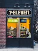 A 7-Eleven store features prominently with a bright yellow ATM sign visible through the window. Inside, various snacks and products are displayed on shelves. The exterior includes a brick wall on the left and a dark panel on the right with overhead lights.
