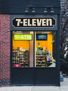 A 7-Eleven store features prominently with a bright yellow ATM sign visible through the window. Inside, various snacks and products are displayed on shelves. The exterior includes a brick wall on the left and a dark panel on the right with overhead lights.
