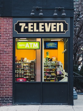 A 7-Eleven store features prominently with a bright yellow ATM sign visible through the window. Inside, various snacks and products are displayed on shelves. The exterior includes a brick wall on the left and a dark panel on the right with overhead lights.