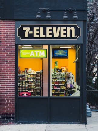 A 7-Eleven store features prominently with a bright yellow ATM sign visible through the window. Inside, various snacks and products are displayed on shelves. The exterior includes a brick wall on the left and a dark panel on the right with overhead lights.