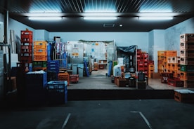 A storage room filled with stacks of colorful plastic crates and boxes organized on the floor and shelves. Fluorescent lights illuminate the space, creating a slightly industrial atmosphere. Various items are placed on the pallets and some walls show signs of wear.