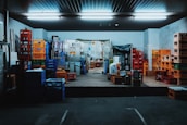 A storage room filled with stacks of colorful plastic crates and boxes organized on the floor and shelves. Fluorescent lights illuminate the space, creating a slightly industrial atmosphere. Various items are placed on the pallets and some walls show signs of wear.