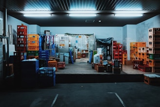 A storage room filled with stacks of colorful plastic crates and boxes organized on the floor and shelves. Fluorescent lights illuminate the space, creating a slightly industrial atmosphere. Various items are placed on the pallets and some walls show signs of wear.