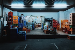 A storage room filled with stacks of colorful plastic crates and boxes organized on the floor and shelves. Fluorescent lights illuminate the space, creating a slightly industrial atmosphere. Various items are placed on the pallets and some walls show signs of wear.