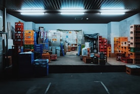 A storage room filled with stacks of colorful plastic crates and boxes organized on the floor and shelves. Fluorescent lights illuminate the space, creating a slightly industrial atmosphere. Various items are placed on the pallets and some walls show signs of wear.