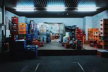 A storage room filled with stacks of colorful plastic crates and boxes organized on the floor and shelves. Fluorescent lights illuminate the space, creating a slightly industrial atmosphere. Various items are placed on the pallets and some walls show signs of wear.