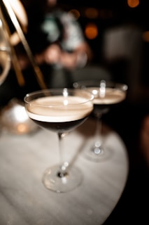 Two cocktail glasses filled with a dark liquid and frothy top are placed on a marble table. The background is dimly lit and slightly blurred, suggesting a relaxed or intimate setting.