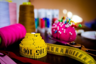 A brightly colored and neatly coiled yellow measuring tape sits on a table alongside sewing supplies. Visible are vibrant spools of thread in brown, pink, and other colors, as well as a pink pin cushion adorned with colorful pins. The background features more thread spools, slightly out of focus, creating a busy but organized craft scene.