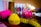 A brightly colored and neatly coiled yellow measuring tape sits on a table alongside sewing supplies. Visible are vibrant spools of thread in brown, pink, and other colors, as well as a pink pin cushion adorned with colorful pins. The background features more thread spools, slightly out of focus, creating a busy but organized craft scene.