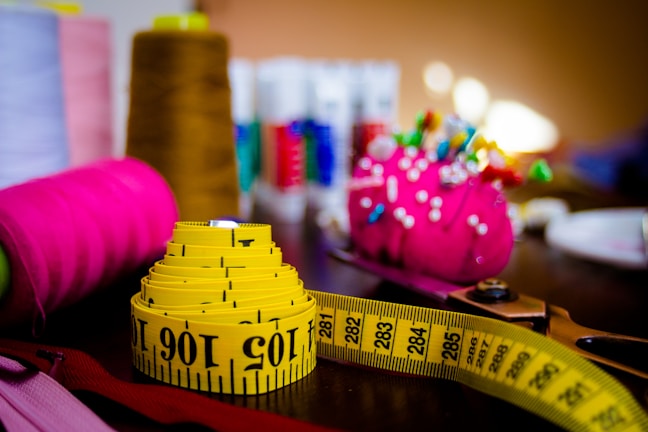 A brightly colored and neatly coiled yellow measuring tape sits on a table alongside sewing supplies. Visible are vibrant spools of thread in brown, pink, and other colors, as well as a pink pin cushion adorned with colorful pins. The background features more thread spools, slightly out of focus, creating a busy but organized craft scene.