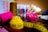 A brightly colored and neatly coiled yellow measuring tape sits on a table alongside sewing supplies. Visible are vibrant spools of thread in brown, pink, and other colors, as well as a pink pin cushion adorned with colorful pins. The background features more thread spools, slightly out of focus, creating a busy but organized craft scene.