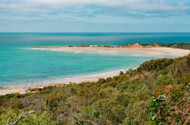 A panoramic view of a pristine beach with turquoise blue waters meeting the sandy shoreline. Lush greenery and shrubs cover the foreground, while gentle waves lap against the shore. The horizon features a gradient of blues in the sky, with a few wispy clouds scattered above.