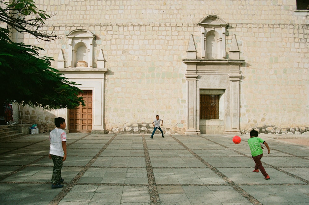 três crianças brincando com bola vermelha perto do edifício durante o dia