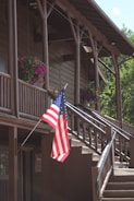 A wooden building with a rustic design features two levels and a sloped roof supported by log beams. Potted pink and purple flowers are hanging along the balcony. An American flag is prominently displayed on a pole near the stairs, which lead to the upper level. Sunlight illuminates parts of the facade, enhancing the natural wood tones.
