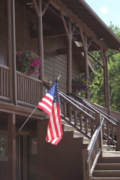 A wooden building with a rustic design features two levels and a sloped roof supported by log beams. Potted pink and purple flowers are hanging along the balcony. An American flag is prominently displayed on a pole near the stairs, which lead to the upper level. Sunlight illuminates parts of the facade, enhancing the natural wood tones.