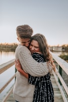 A young couple embraces affectionately on a wooden walkway by a body of water. The man wears a knitted light sweater while the woman is dressed in a patterned dark dress. Their expressions reflect warmth and happiness, and the lighting suggests a sunset or sunrise setting.