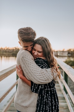 A young couple embraces affectionately on a wooden walkway by a body of water. The man wears a knitted light sweater while the woman is dressed in a patterned dark dress. Their expressions reflect warmth and happiness, and the lighting suggests a sunset or sunrise setting.
