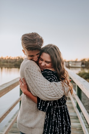 A young couple embraces affectionately on a wooden walkway by a body of water. The man wears a knitted light sweater while the woman is dressed in a patterned dark dress. Their expressions reflect warmth and happiness, and the lighting suggests a sunset or sunrise setting.