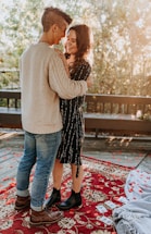 A couple stands close together, smiling at each other on a wooden deck surrounded by trees. Red and orange leaves are scattered on the ground along with some photographs, adding a touch of romance. They are standing on a decorative red rug with floral patterns, and the natural light creates a warm, intimate atmosphere.