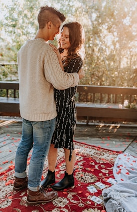 A couple stands close together, smiling at each other on a wooden deck surrounded by trees. Red and orange leaves are scattered on the ground along with some photographs, adding a touch of romance. They are standing on a decorative red rug with floral patterns, and the natural light creates a warm, intimate atmosphere.